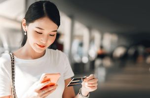 a smiling asian woman looks at her phone and credit card while making an online purchase